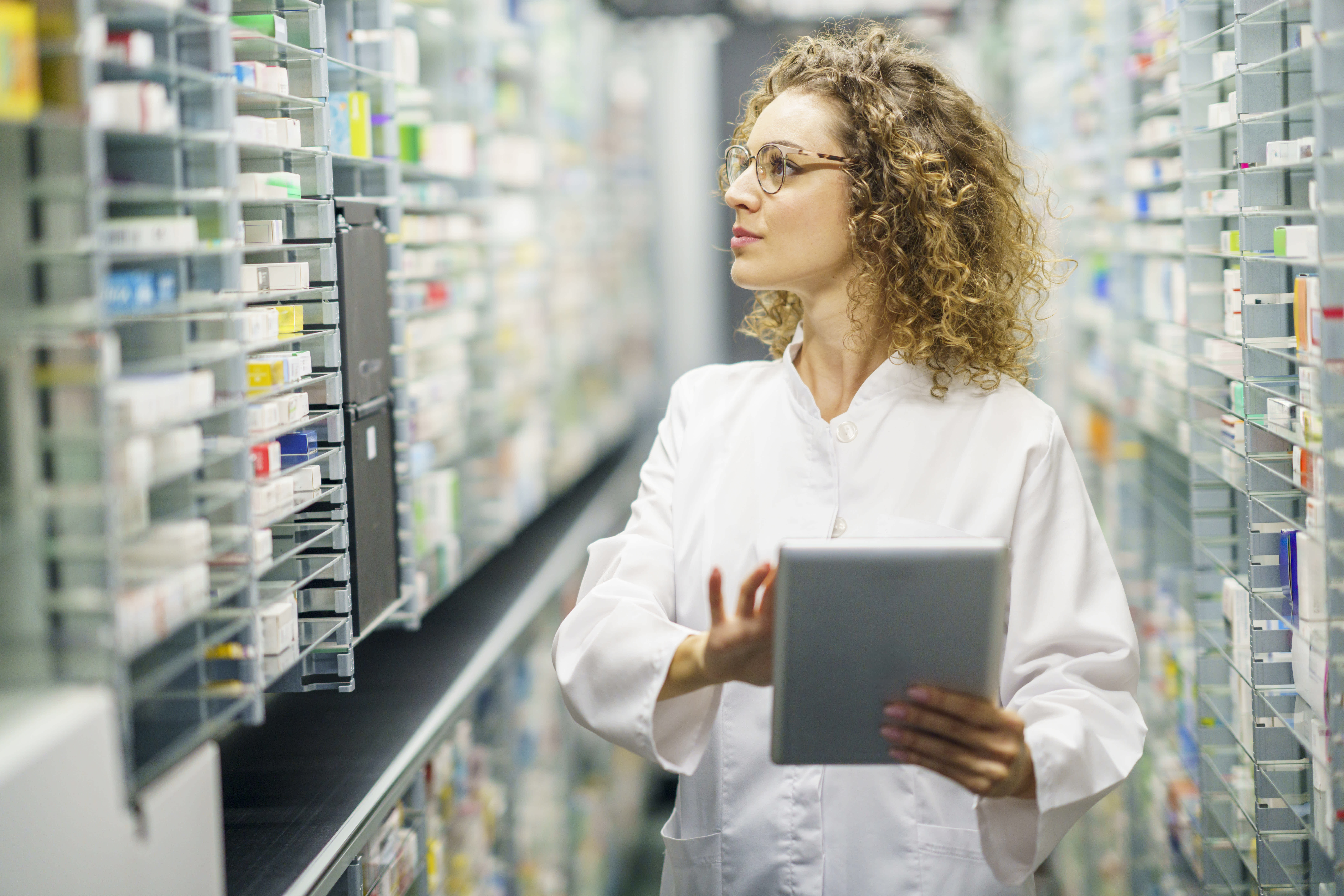Healthcare professional in a white lab coat using a tablet while reviewing medication inventory in a pharmacy aisle with shelves of packaged medicines.