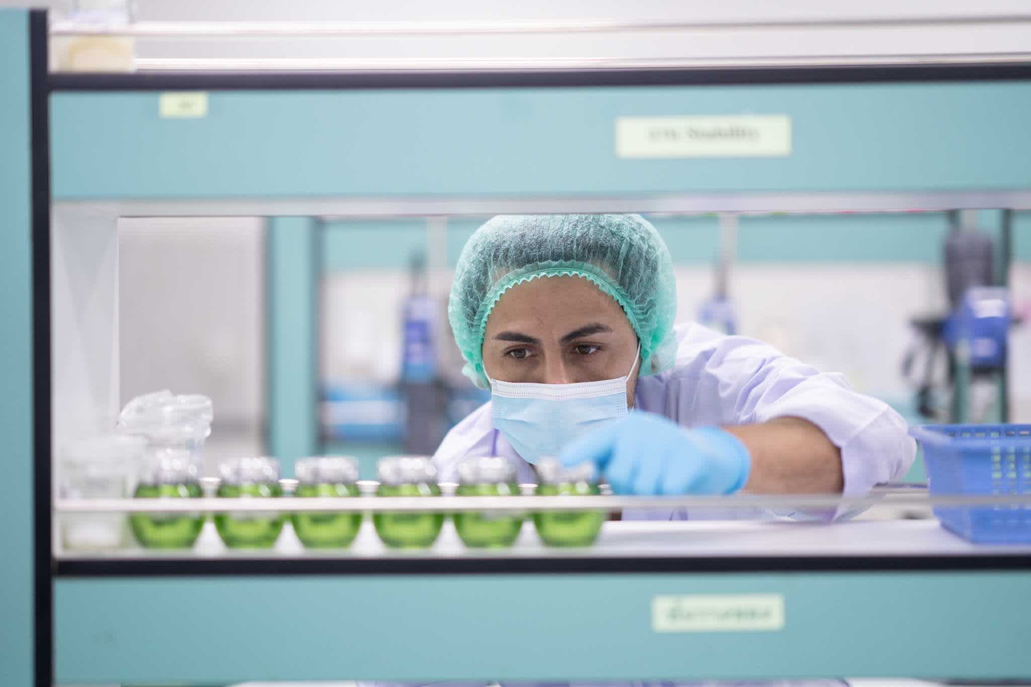 A scientist wearing a mask, gloves, and a hair cover examines small green samples in glass containers on a laboratory shelf.