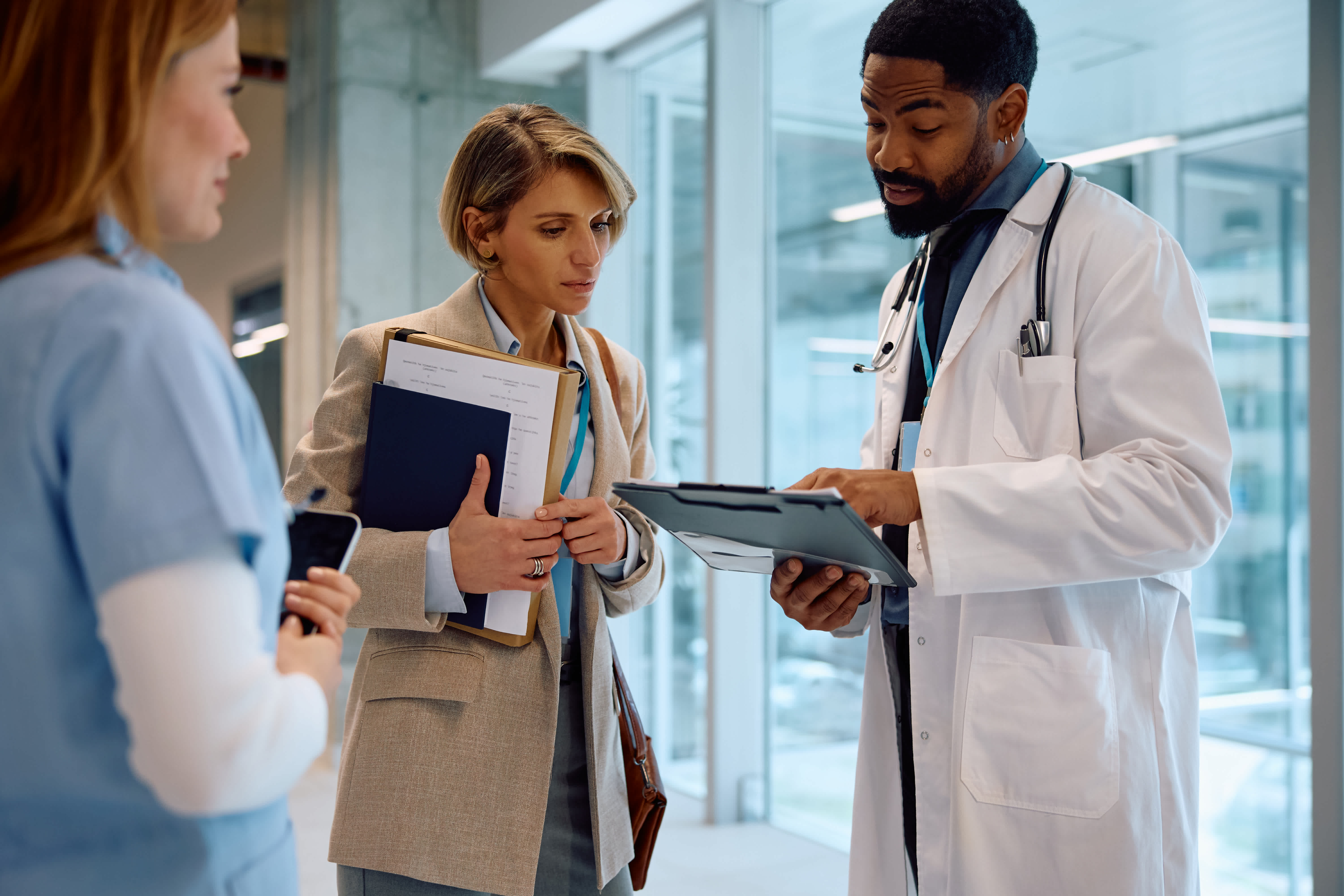A doctor shows information on a tablet to a woman holding folders while another healthcare professional stands nearby in a modern medical facility.