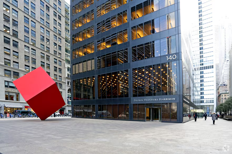A large red cube sculpture stands in front of a modern glass office building with the address 140. The building has many lit windows, and city skyscrapers surround the scene.