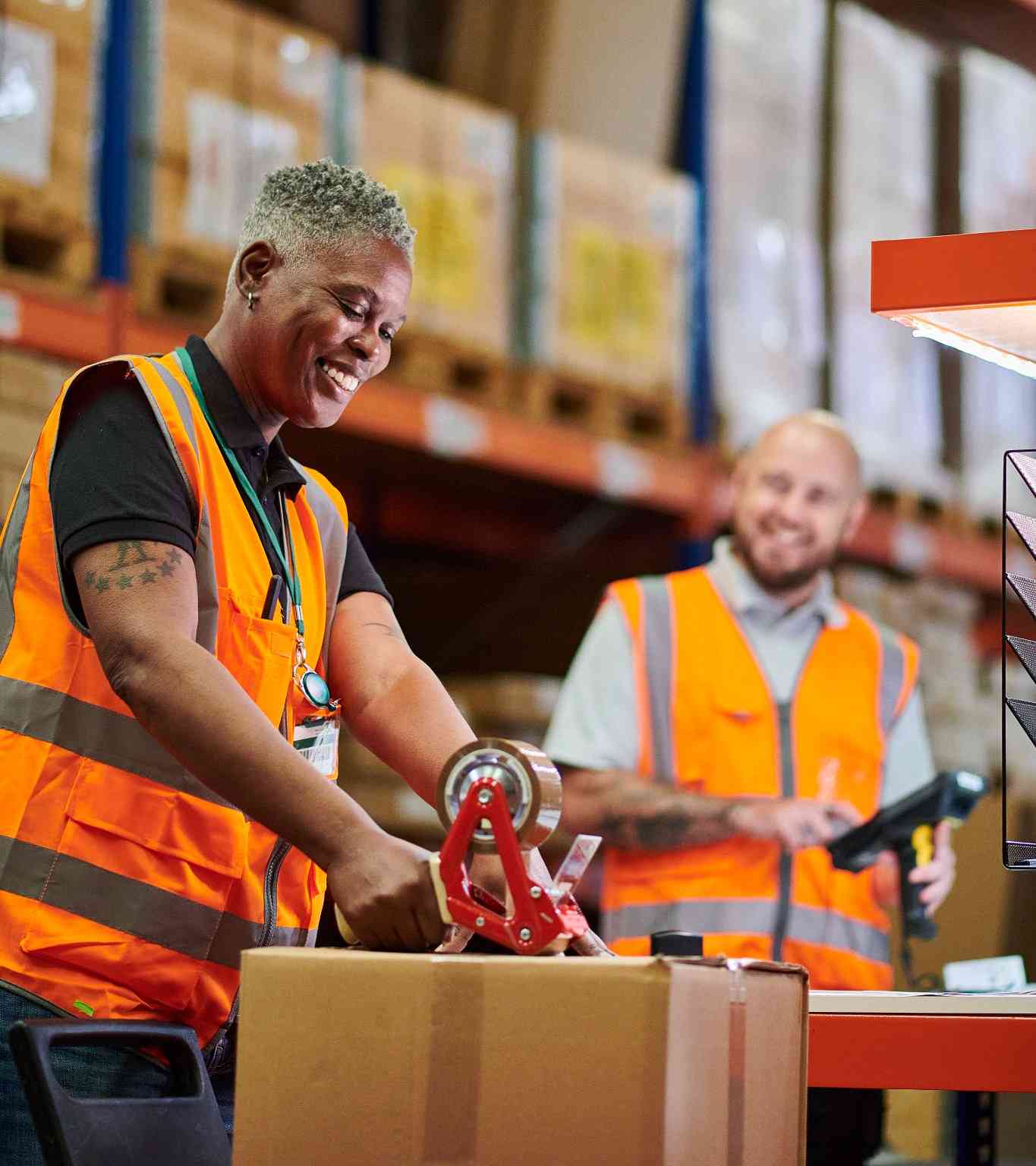 Two workers in orange safety vests pack boxes in a warehouse. One smiles while using packing tape on a box; the other stands nearby holding a handheld scanner, also smiling. Shelves of boxes are visible in the background.
