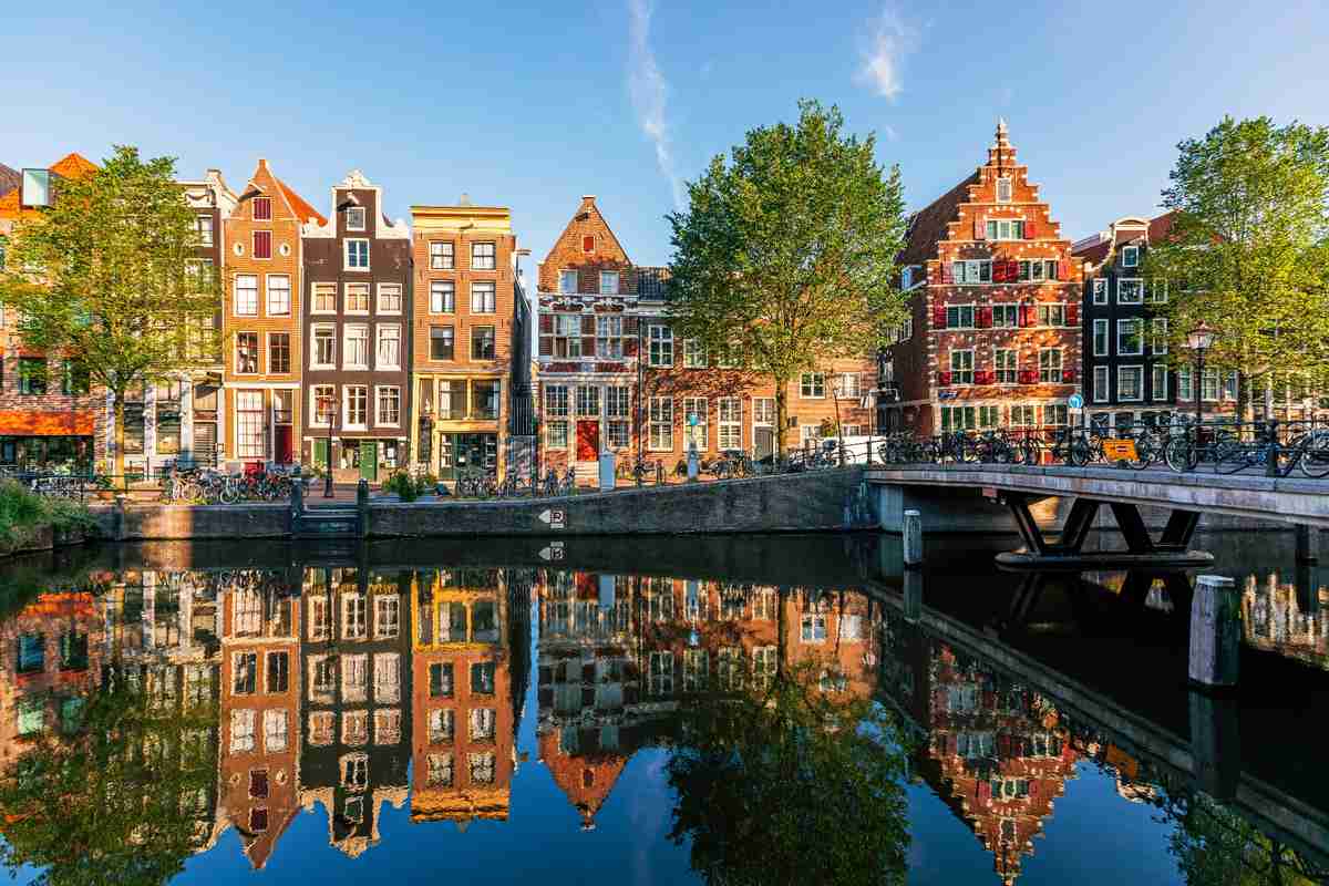 Colorful canal houses and green trees are reflected in the calm water of a canal in Amsterdam, with a bridge and clear blue sky in the background.