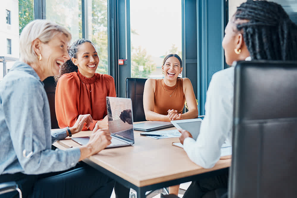 Four women sit around a conference table with laptops and tablets, smiling and talking in a bright, modern office with large windows.
