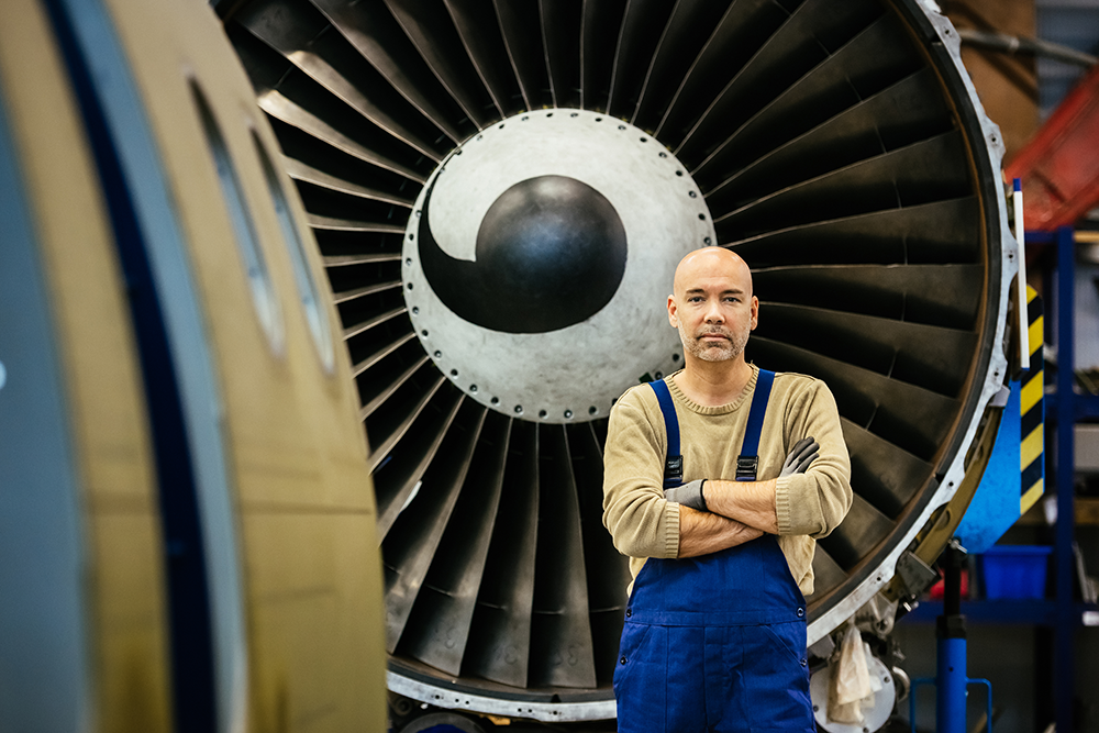 A man in blue overalls stands with arms crossed in front of a large turbine engine inside an industrial facility.