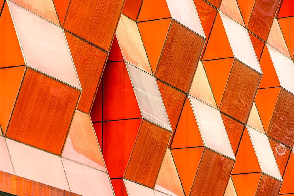 Close-up of an abstract building facade with repeating orange and beige geometric panels arranged in a three-dimensional, zigzag pattern.