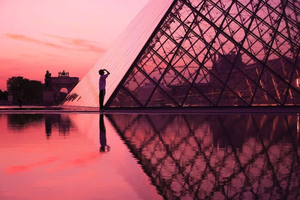 A person stands near the Louvre Pyramid at sunset, silhouetted against the pink sky and glass structure, with reflections in the water.
