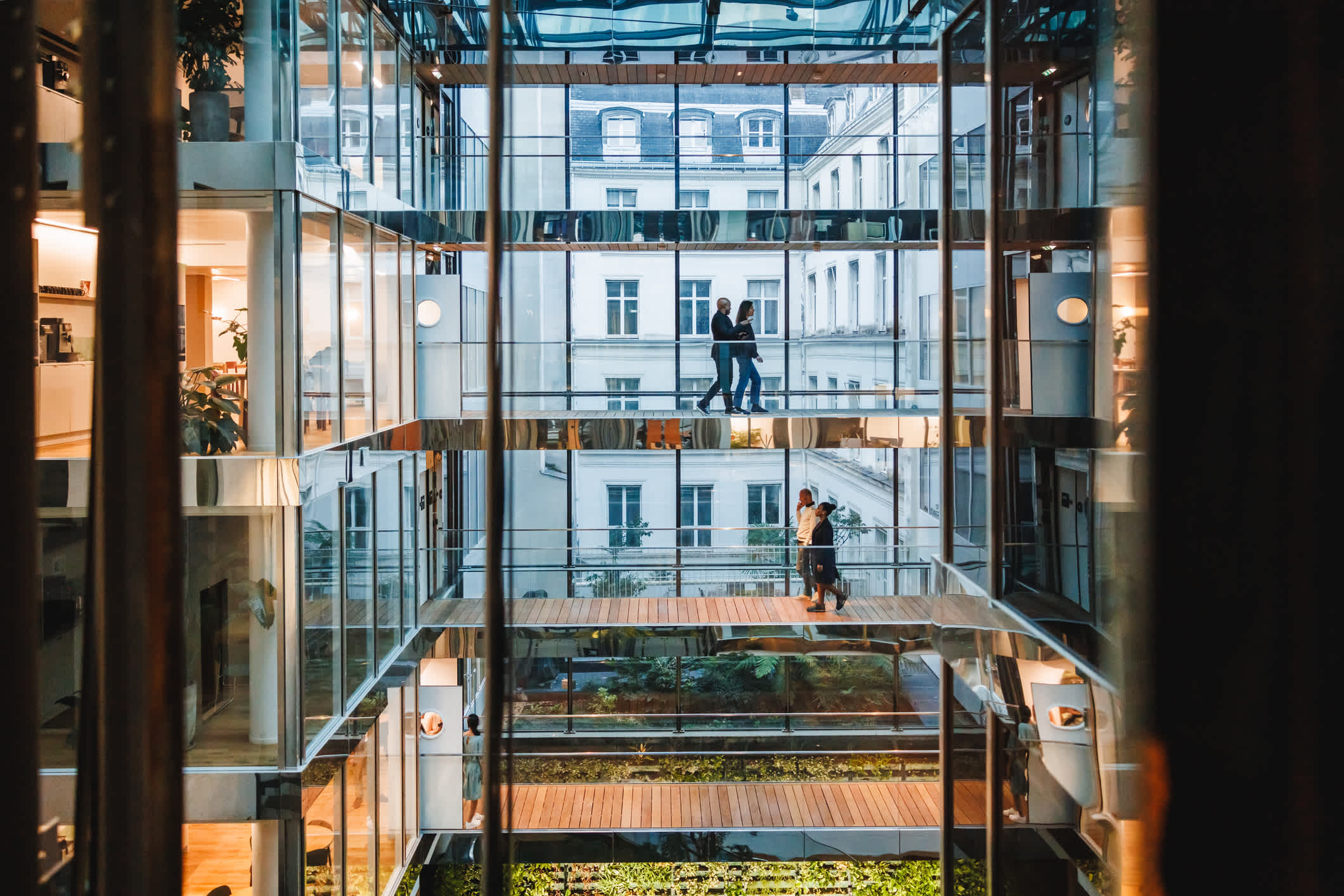 Modern glass atrium with multiple floors, people walking on skywalks, and large windows revealing an exterior building. Natural light fills the space.