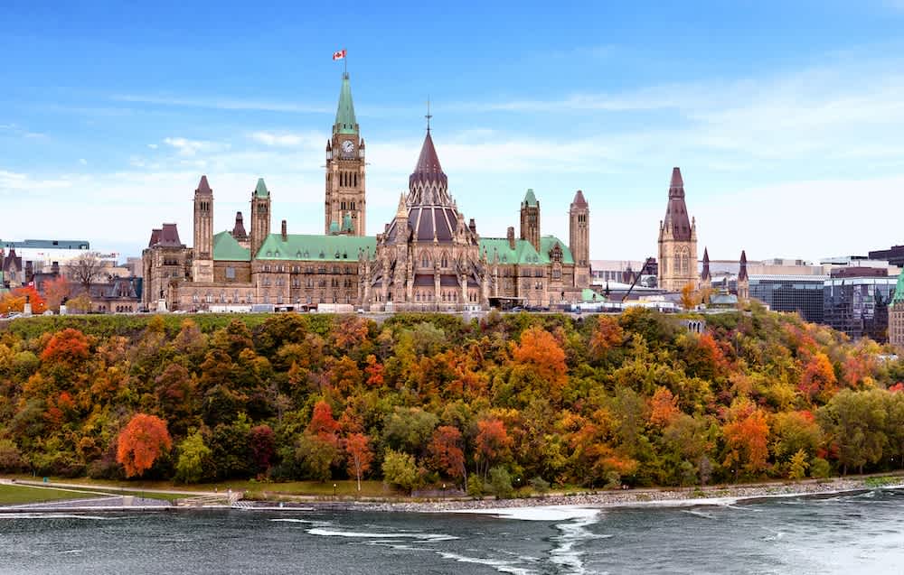 Canadas Parliament buildings in Ottawa are seen atop a hill, surrounded by colorful autumn trees with a river in the foreground and a blue sky above.