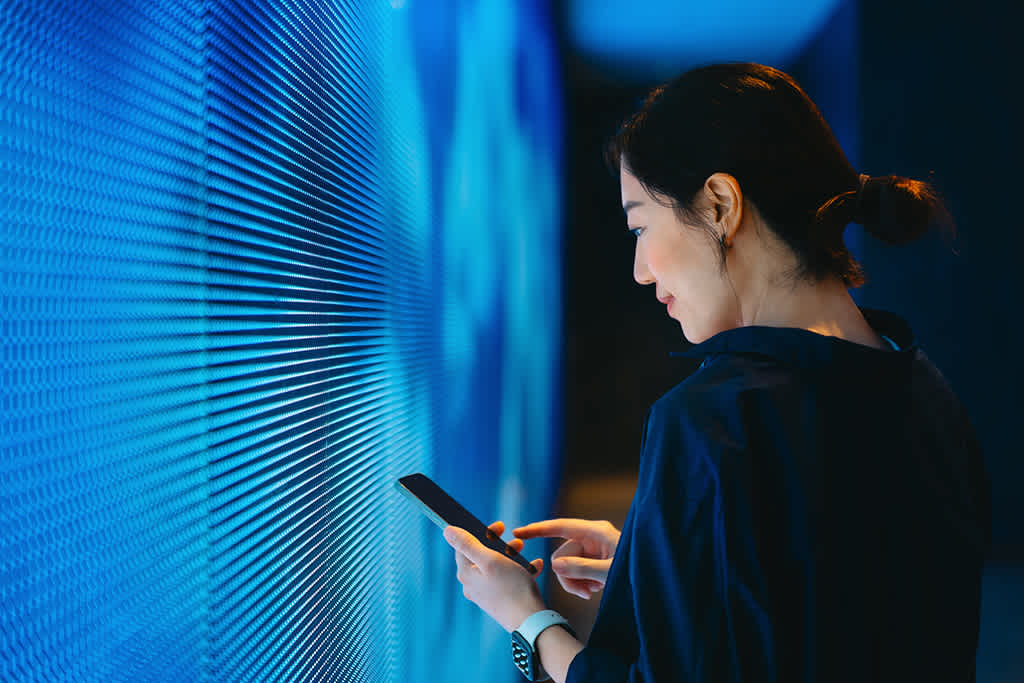 A woman stands indoors near a glowing blue wall, looking at and using her smartphone with both hands. The scene has a modern, tech-inspired atmosphere.