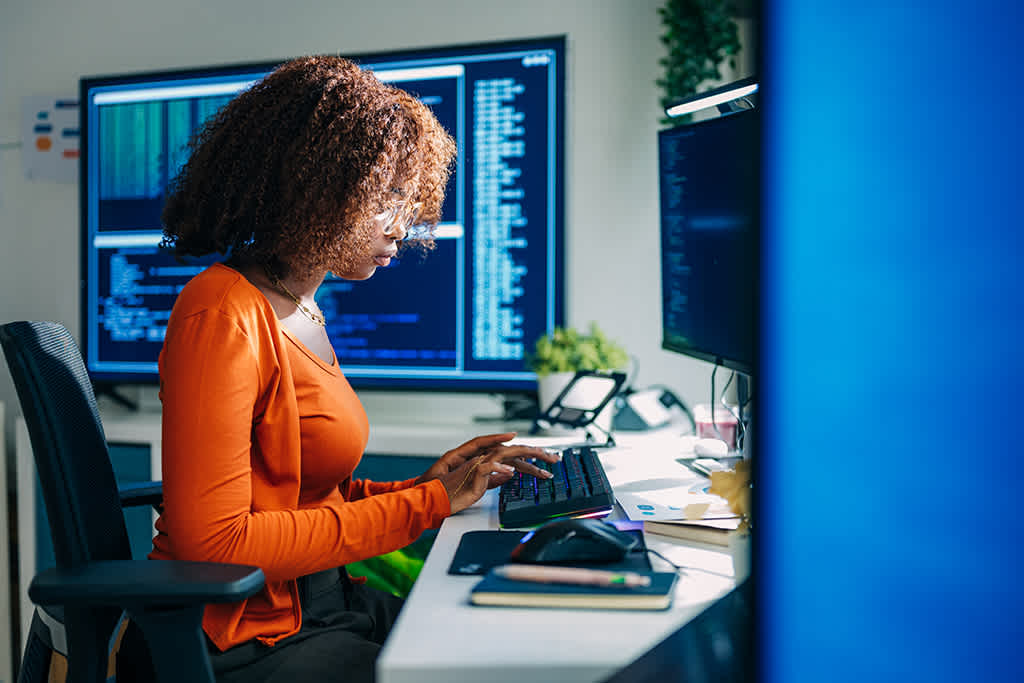 A woman with curly hair in an orange top types on a keyboard at a desk with multiple computer screens displaying code in a modern office setting.