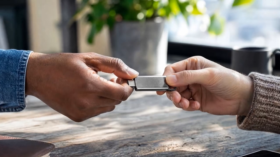 Two people exchange a USB flash drive across a wooden table, with a potted plant blurred in the background.