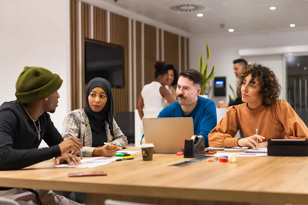 Four people sit around a table in an office, engaged in discussion. They have notebooks, papers, and a laptop. Two more people stand talking in the background. The setting is modern and collaborative.