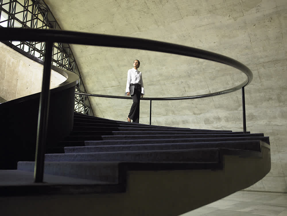 A woman in business attire stands on a modern spiral staircase with a concrete wall and large windows in the background.