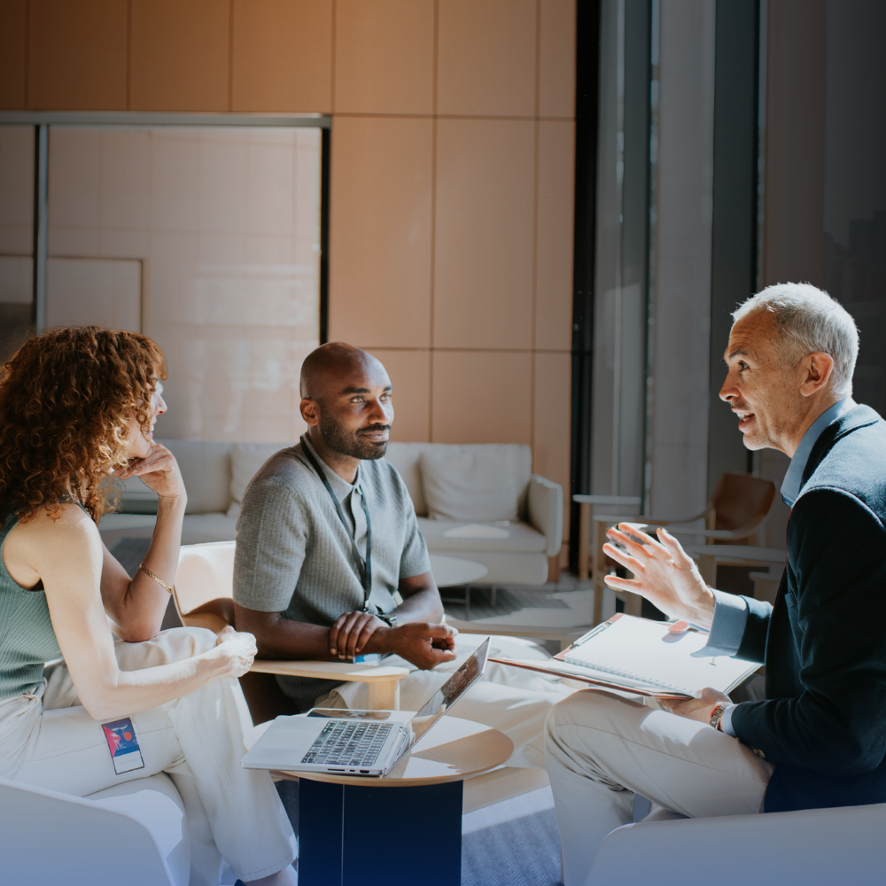 Three people sit in a modern office lounge, engaged in discussion. One person takes notes, another listens attentively, and a laptop is open on the table between them. Natural light fills the space.