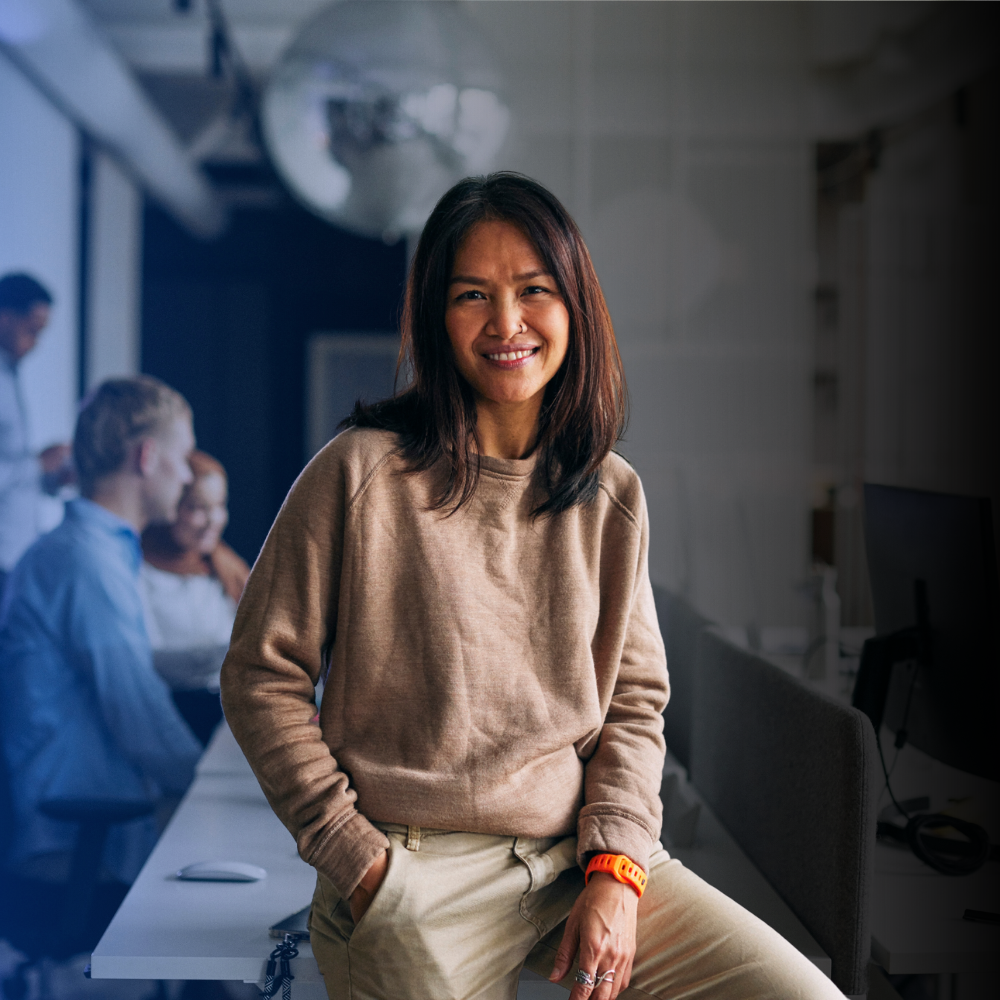 A woman in a beige sweater smiles while sitting on a desk in a modern office, with three people conversing in the blurred background.