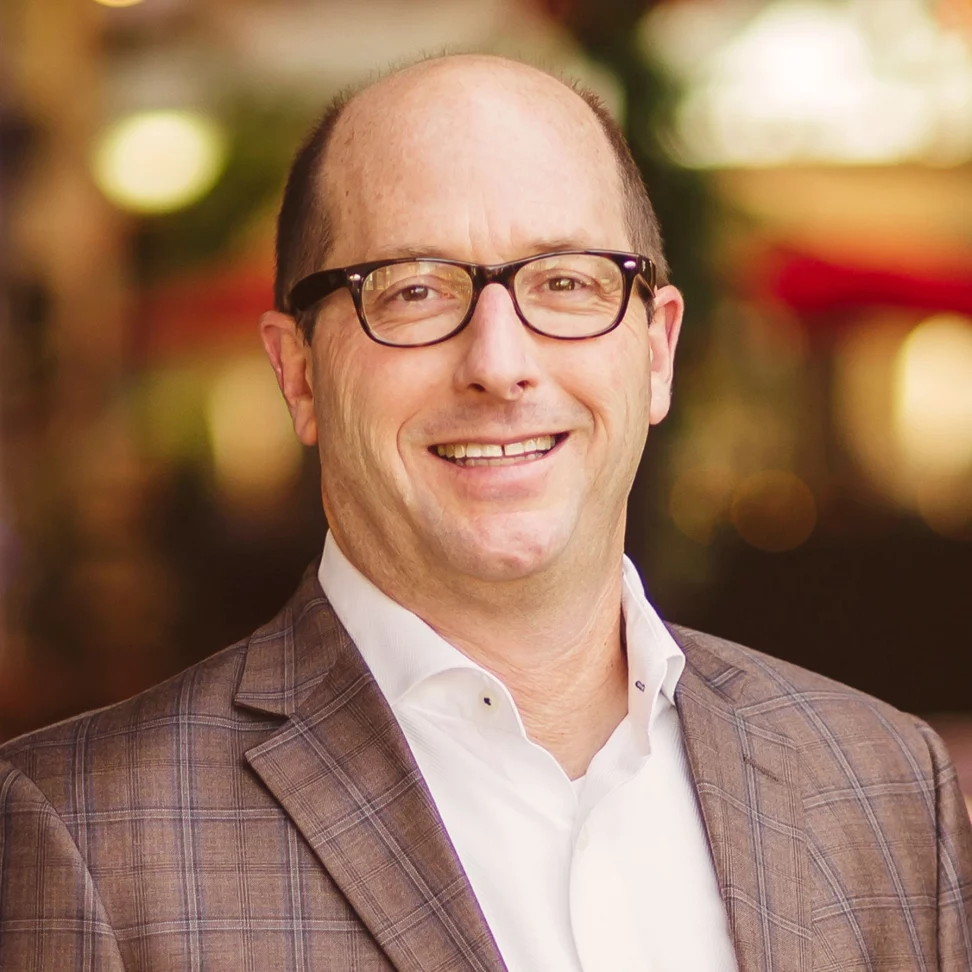 A man wearing glasses, a white shirt, and a plaid suit jacket smiles at the camera. The background is softly blurred with warm lights and hints of greenery.