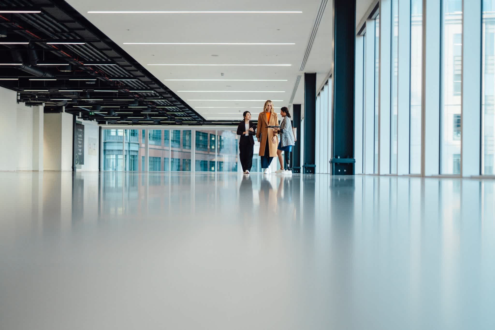Three women in business casual attire walk together down a bright, modern office hallway with floor-to-ceiling windows.