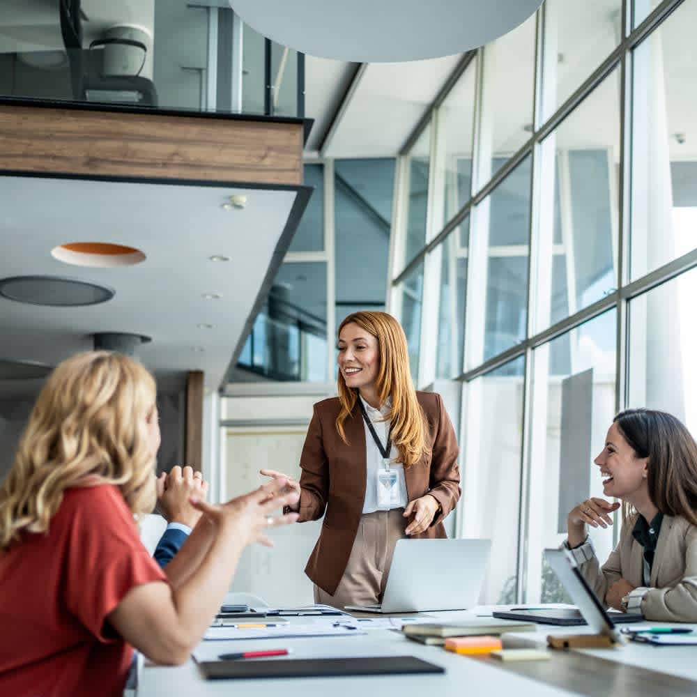 A woman in a brown blazer leads a meeting, speaking to two colleagues seated across a conference table.