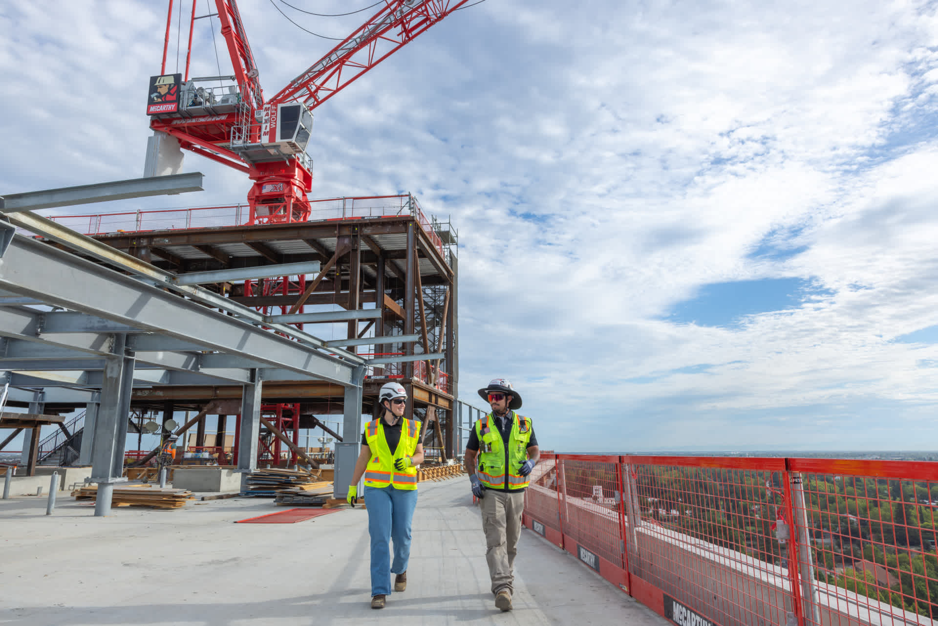Two construction workers in safety vests and hard hats stand facing a multi-story building under construction. One wears an orange vest, the other a yellow one. Scaffolding and part of a crane are visible in the background, under a clear sky.