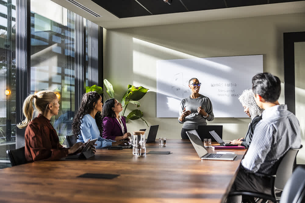A woman stands at the front of a boardroom, presenting to a group of colleagues seated around a large wooden table.