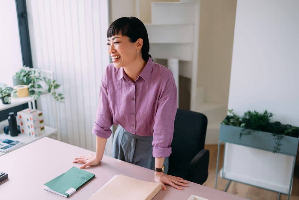 A woman in a purple button-down shirt stands smiling at a desk in a bright office.
