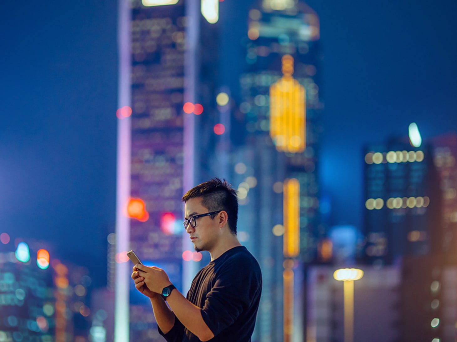 man in glasses, looking at phone in front of city scape at night