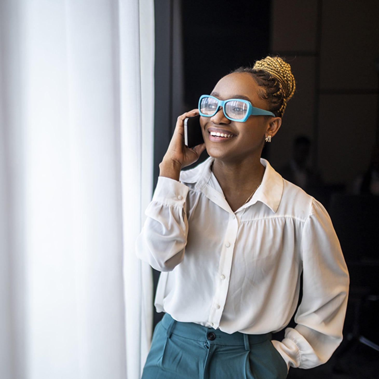 Black woman in teal glasses talking on her phone by a window
