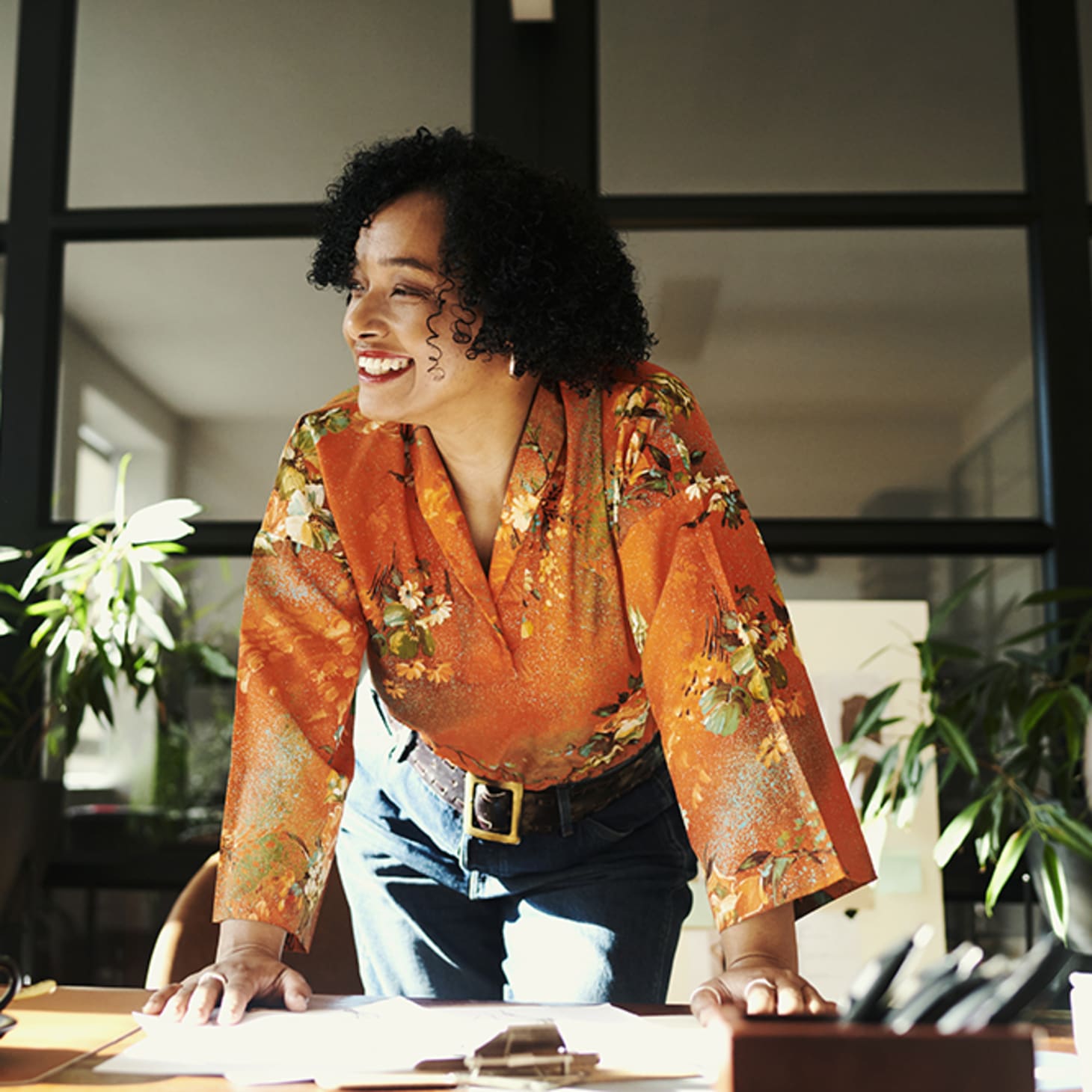 black woman in orange blouse smiling at computer leaning on desk in office with plants