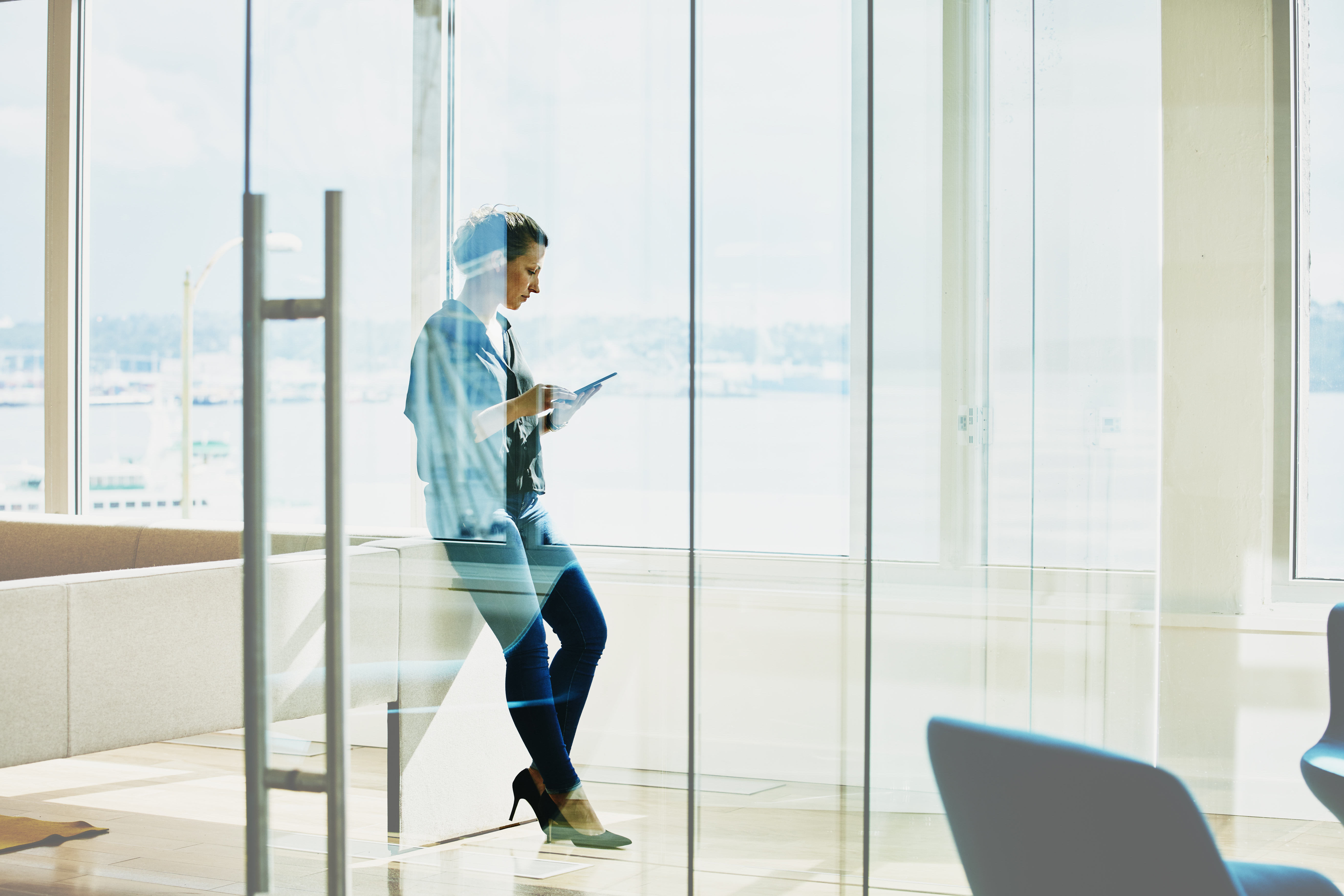 full-length photo of a woman leaning against a wall in a windowed office, looking at her phone