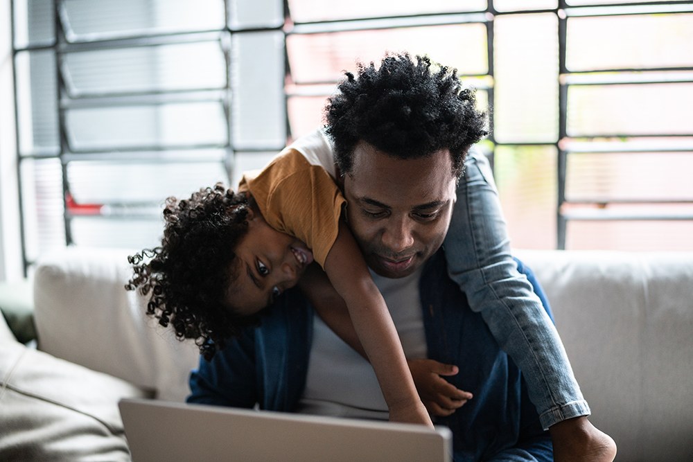 Black man with child on his back, working on his laptop on the couch