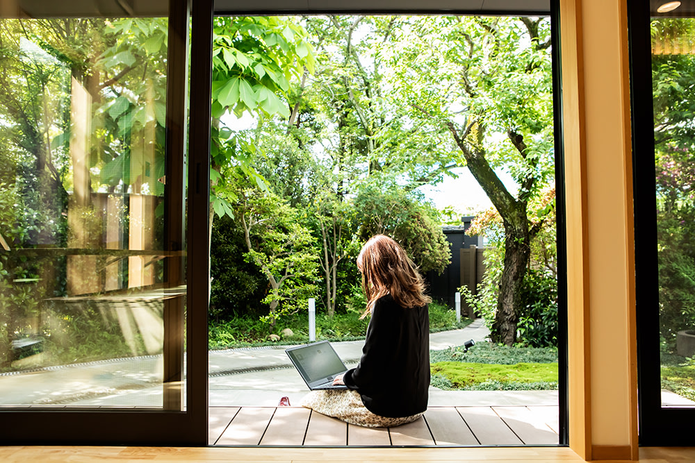looking through sliding door, woman sitting on deck steps working on laptop surrounded by green yard and trees