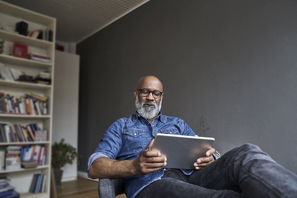 black man with grey beard and glasses looking down reading tablet at home in office with bookshelf