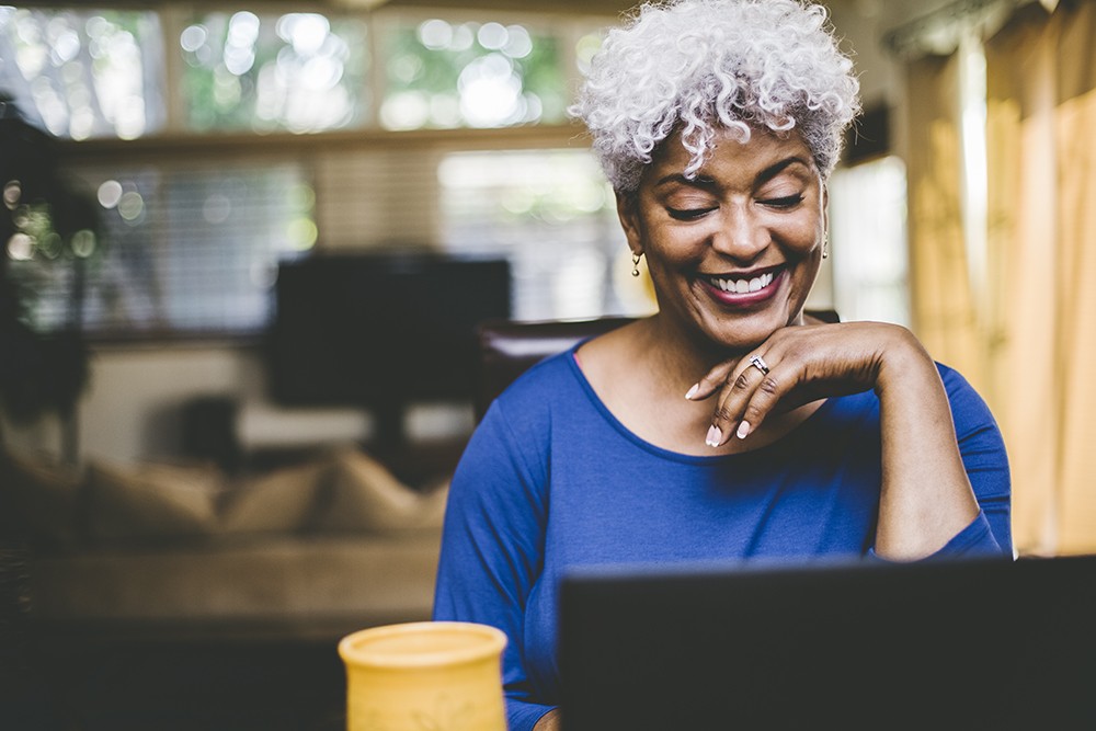 black woman with white and grey hair wearing blue top smiling at laptop sitting at desk