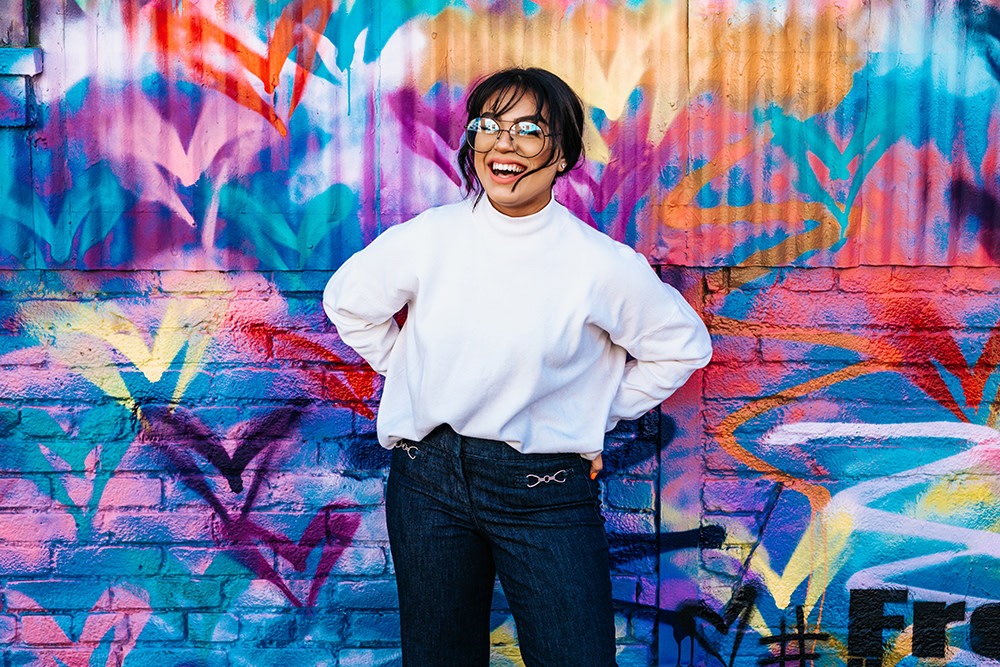 A woman smiling stands in front of a vibrant, graffiti-covered wall. She wear glasses, a white top, and jeans. The wall features colorful heart and swirl patterns in shades of pink, blue, and orange.