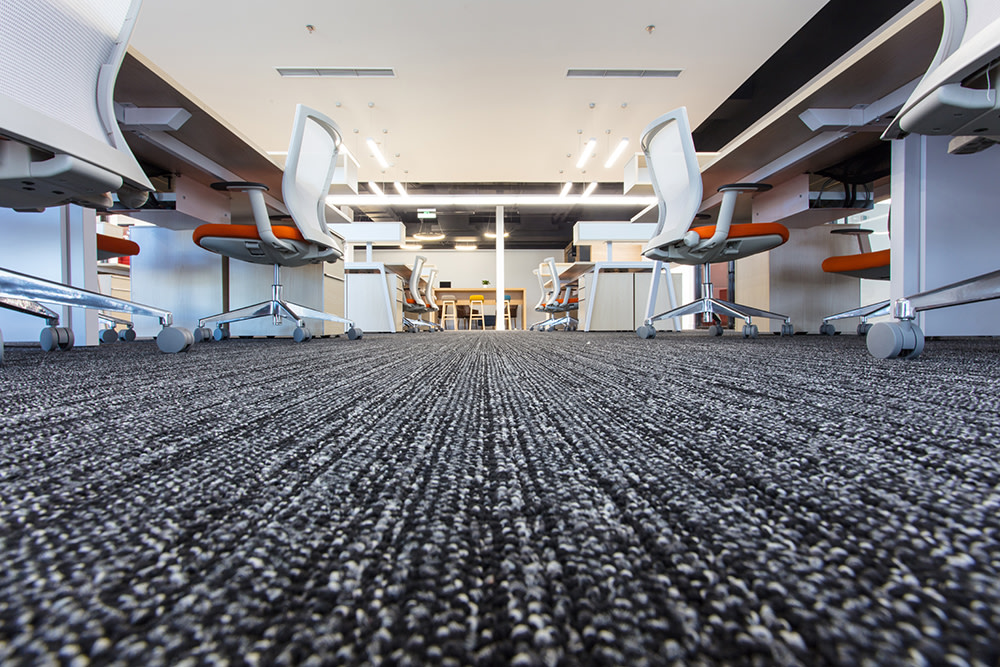 Low, ground level view of carpeted, minimalist, empty office, down an aisle of white desks with orange-seated chairs