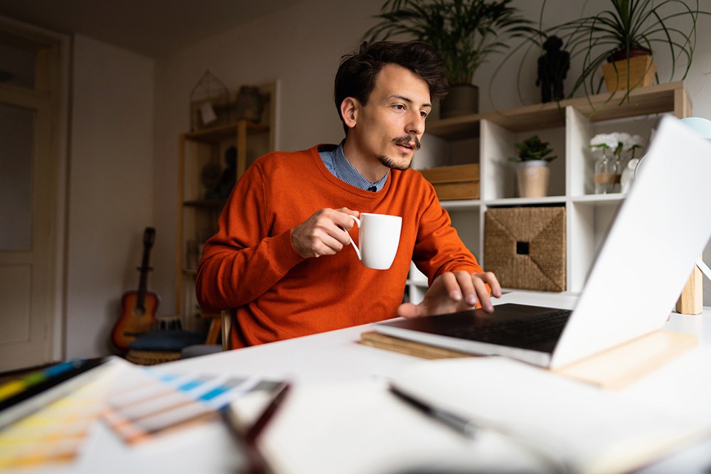 white man with mustache and goatee in orange shirt, drinking coffee while working on laptop