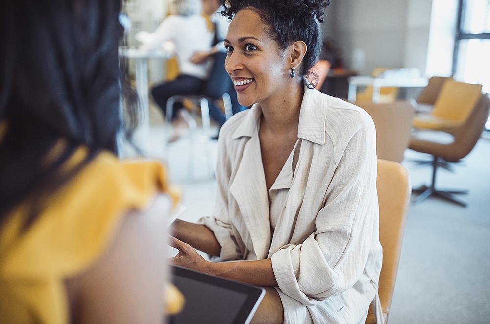 Black woman in white jacket looking happily shocked in discussion with colleauge