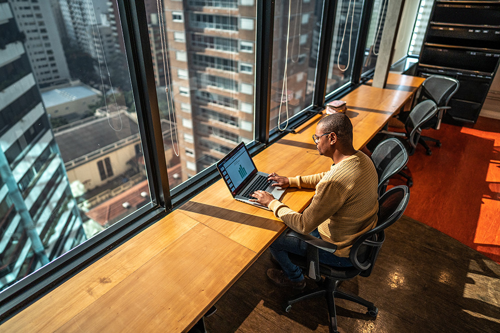 black man sitting at long row desk typing on laptop overlooking city buildings below