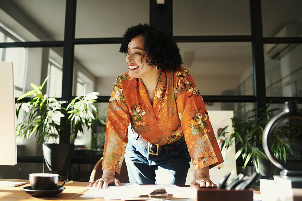 A woman in an orange floral blouse leans forward on a desk, smiling and looking away. She is in a bright office with potted plants and large windows. A cup sits on the desk beside her.