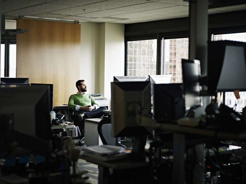 Man in green shirt sitting along in empty office looking out the window at city buildings