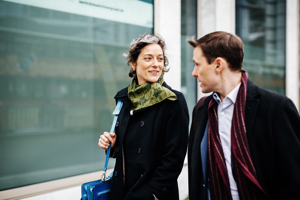 Two people walking and talking outside. One is wearing a black coat and green scarf, the other a dark jacket with a maroon scarf. They are on a sidewalk in front of a window.