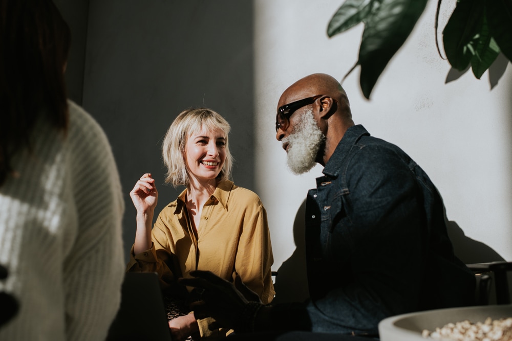 A woman with short blonde hair and a man with a white beard and sunglasses are sitting and talking in a sunlit room. The woman is smiling and wearing a yellow blouse. Green plants are visible in the background.