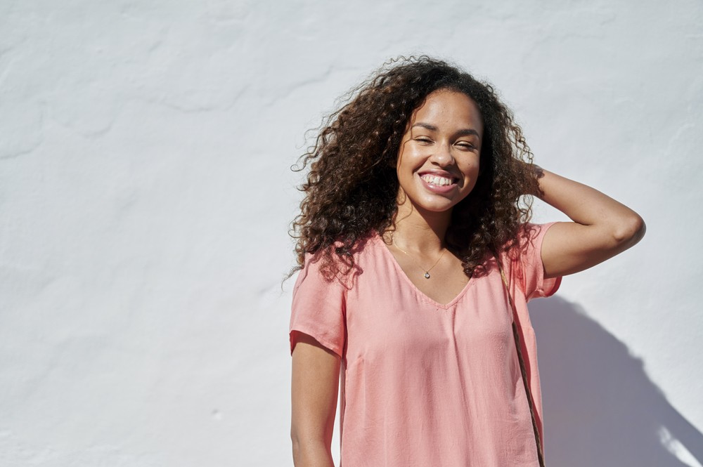 black woman with hair down smiling wearing pink shirt standing in the sun in front of white wall