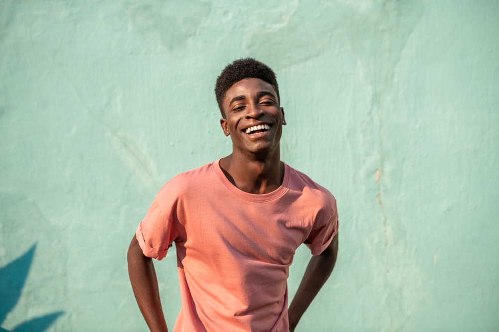 smiling Black man in orange shirt against a teal stucco wall