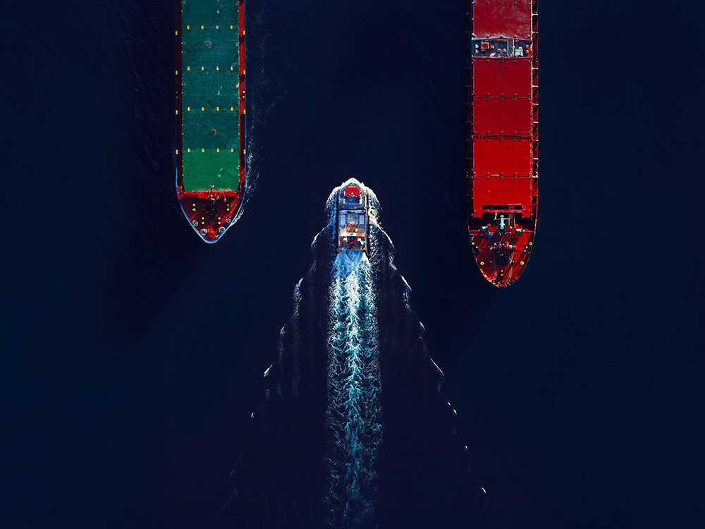 Aerial view of a small boat traveling between two large cargo ships. The ship on the left is green and the one on the right is red. The deep blue water creates a strong contrast with the vibrant colors of the vessels.