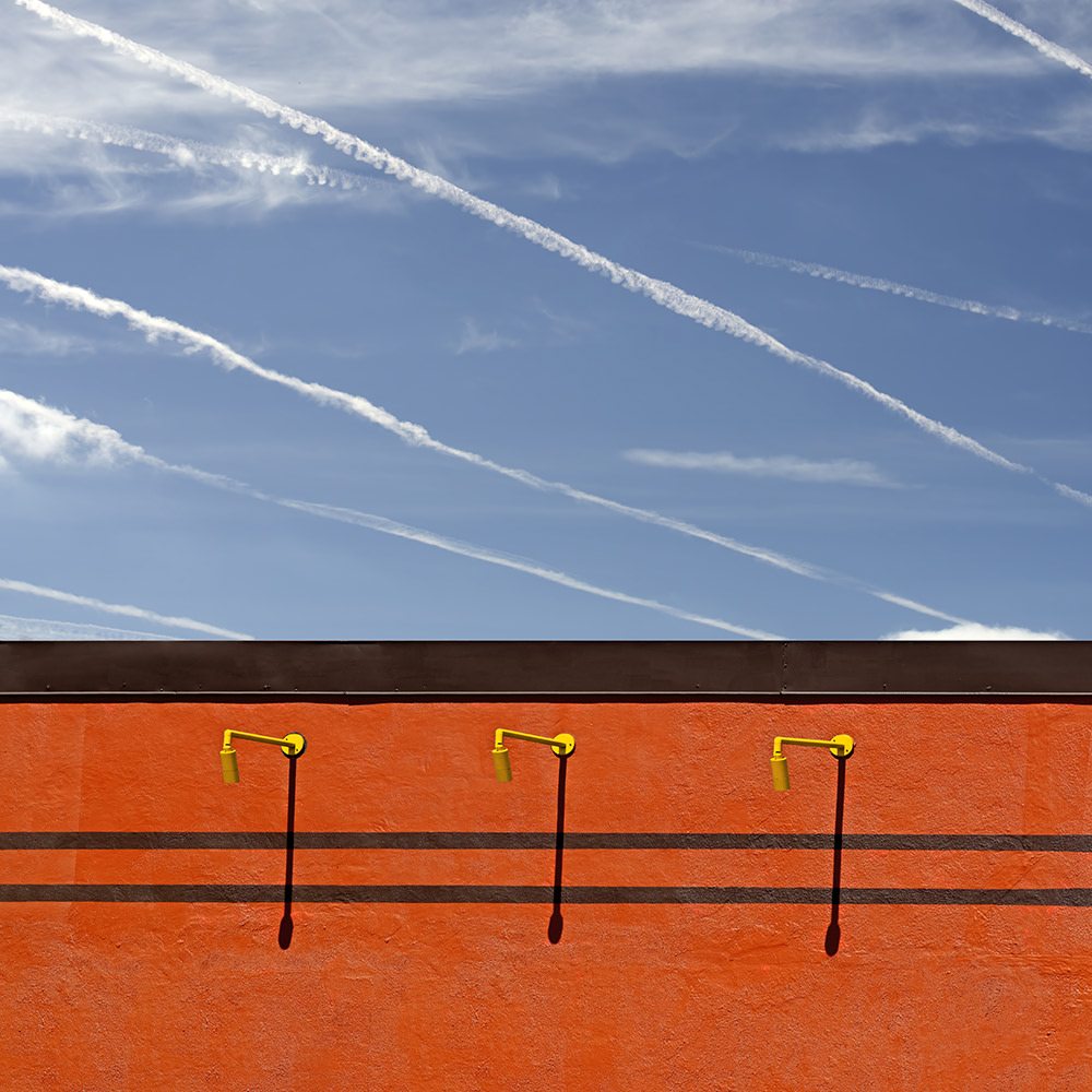 Three yellow spigots protrude from an orange wall with two thin black horizontal lines. The background is a blue sky filled with multiple white contrails and a few wispy clouds.