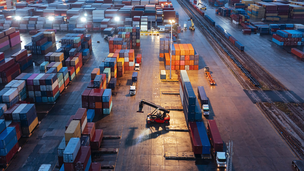 Aerial view of a busy shipping container yard at dusk. Stacks of colorful containers are arranged on either side of the yard. A crane is actively moving a container, with several trucks and vehicles positioned throughout the area.