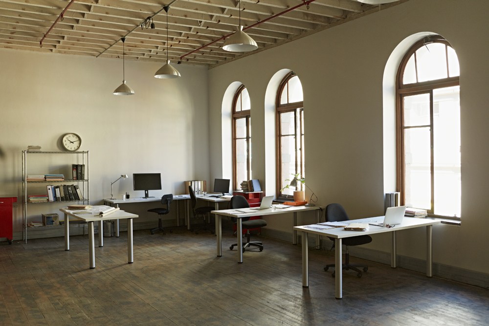 Spacious office with high ceilings, featuring large arched windows. Four desks are arranged with computers, books, and chairs. A metal shelf holds various items, and a wall clock is visible. Natural light fills the room.