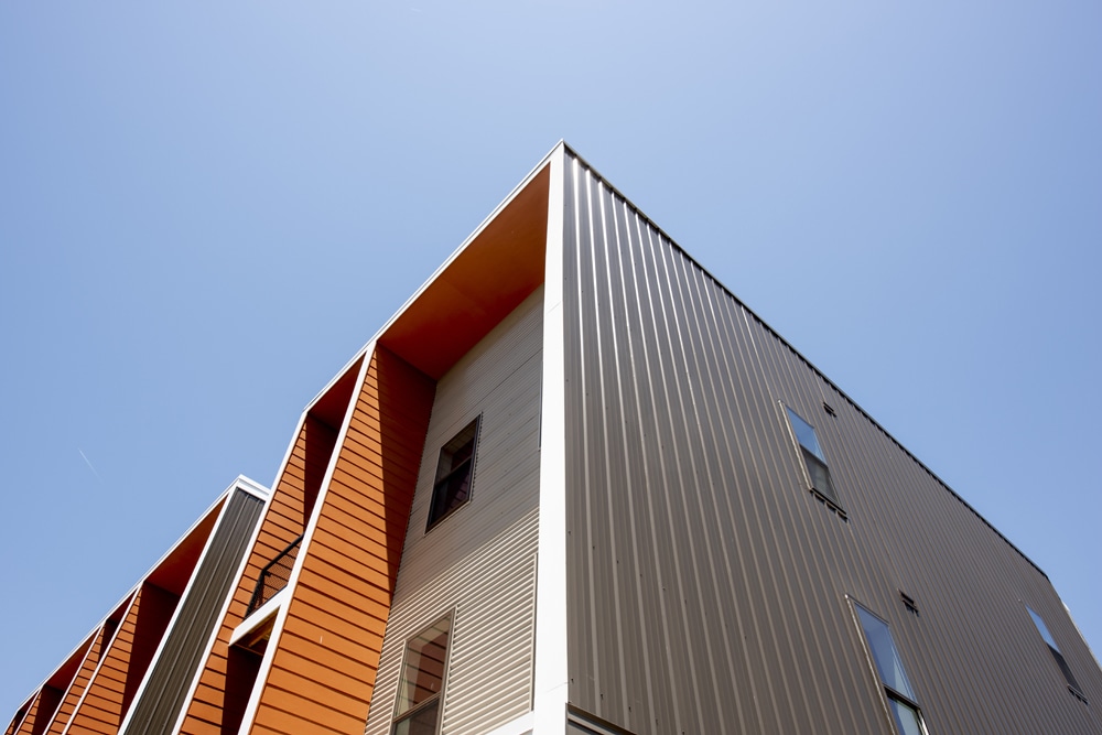 A modern building with sharp lines features a mix of orange and gray vertical panels against a clear blue sky. The building has multiple window openings and a contemporary architectural design. 