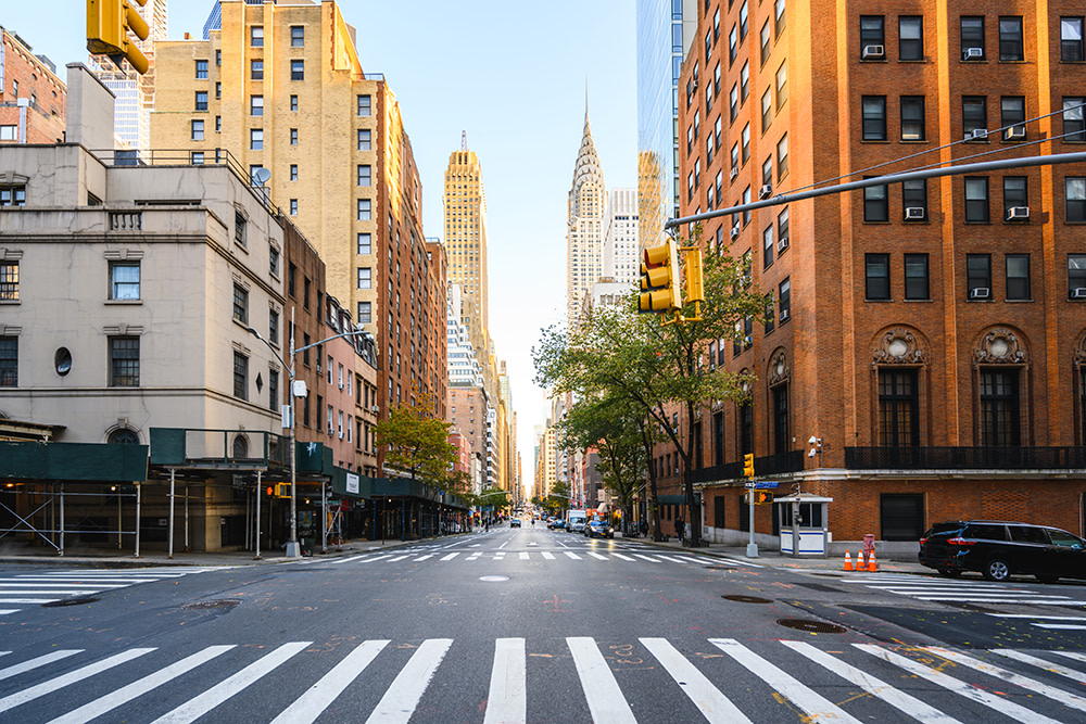 A city street with a wide crosswalk and lined with tall buildings, including some with brick facades. The iconic Chrysler Building is visible in the background. There are traffic lights, trees, and a clear blue sky.